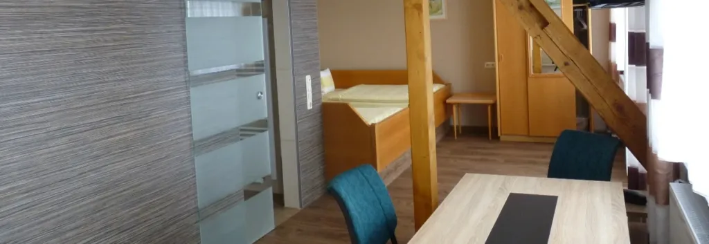 Apartment mit Bett, Schiebetür zum Bad, Esstisch mit Stühlen und Holztreppe