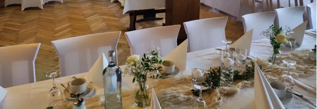 Hochzeitstafel mit weißen Stuhlhussen, Blumengestecken und Parkettboden im großen Gastzimmer