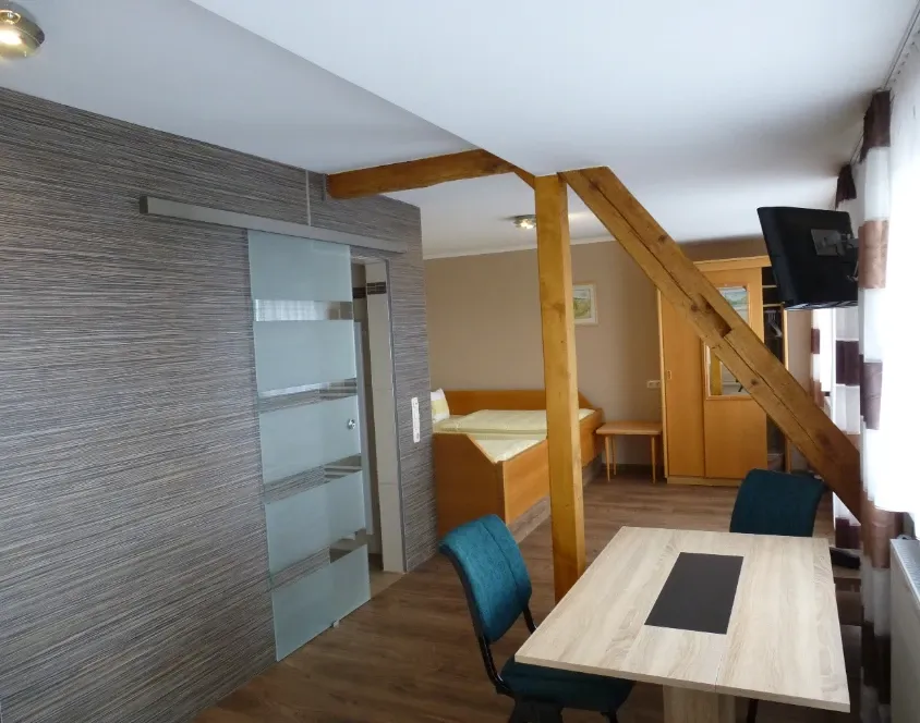 Apartment mit Bett, Schiebetür zum Bad, Esstisch mit Stühlen und Holzbalkendecke