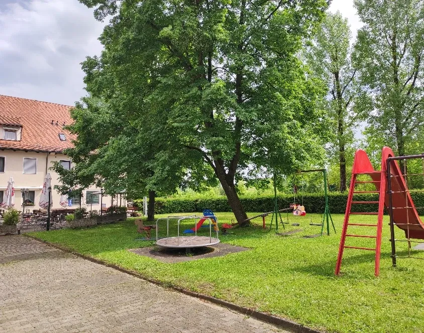 Spielplatz neben dem Hotel mit roter Rutsche und Schaukel unter großen Bäumen