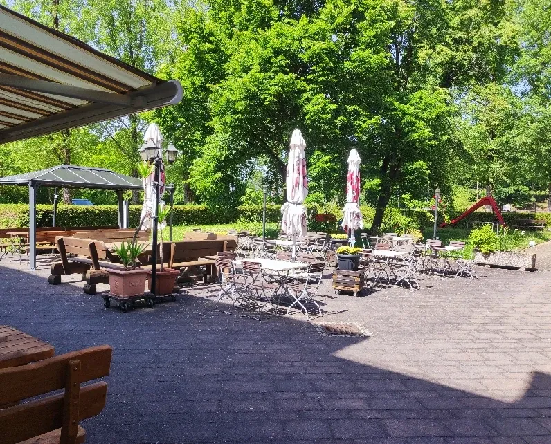 Biergarten mit Bistrotischen, Stühlen und weißen Sonnenschirmen unter Bäumen